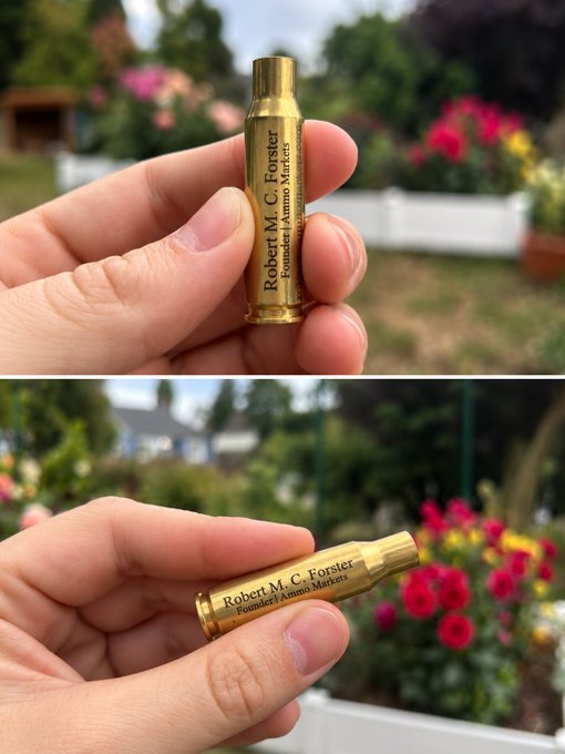Two views of a hand holding a small, gold, bullet-shaped object engraved with 'Robert M. C. Forster, Founder | Ammo Markets' against a background of flowers.