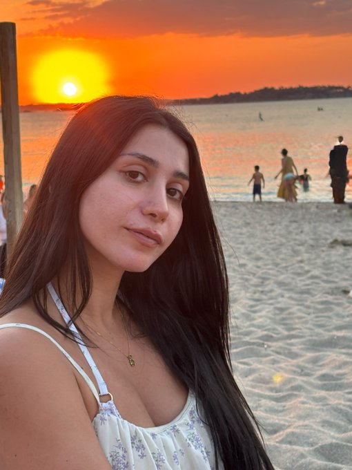 A selfie of a young woman with long dark hair at a beach during sunset, wearing a white floral top with a gold necklace.