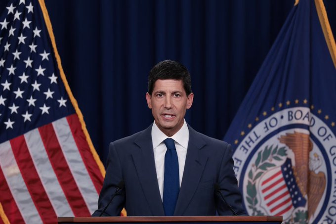A man in a dark suit and blue tie speaks at a podium in front of an American flag and the seal of the Board of Governors of the Federal Reserve.