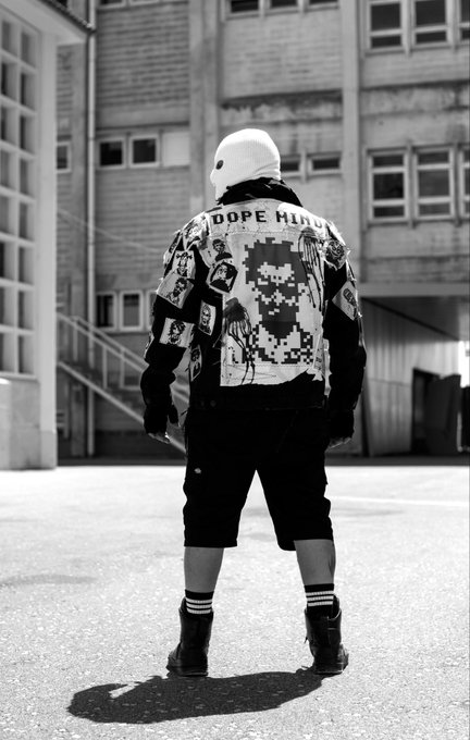 A black and white photo of a person standing in an urban courtyard wearing a graphic jacket with 'DOPE HIDU' text, a pixelated character design on the back, shorts, and a mask.