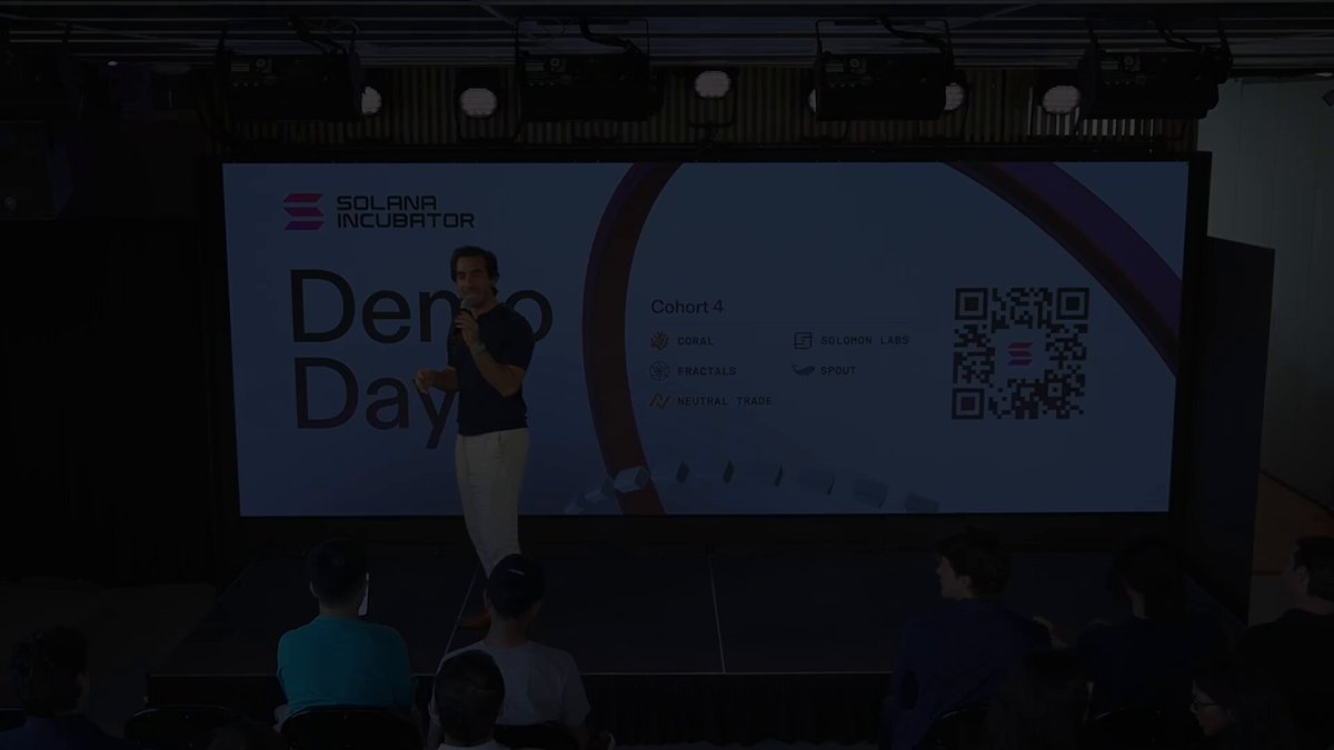 The image shows a room with a presentation screen featuring an image of people standing behind a podium with a microphone, likely presenting or giving a speech at a "Cohort 4 Demo Day". The attendees include people sitting on chairs surrounding the table in front of them. A large screen displays text related to the event, which is about a technology showcase for apps developed by teams from Solana, one of several companies participating. A Q&A session seems to be happening with an audience member asking questions at this demonstration event.