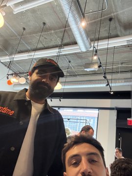 A selfie of a bearded man wearing a black cap and a black button-up shirt over a white t-shirt, standing in a modern indoor space with exposed ceilings and hanging lights.