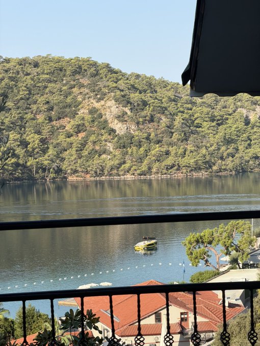 A scenic view from a balcony overlooking a calm bay or inlet with forested hills, showing a yellow boat on the water and houses with terracotta roofs along the shoreline.