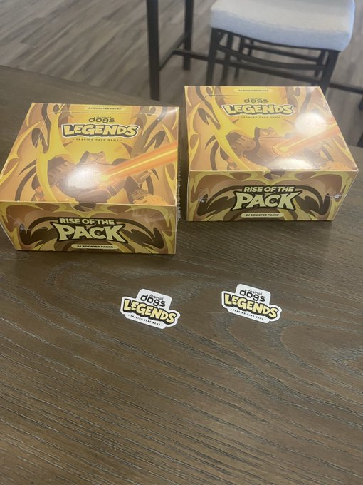Two yellow 'Rise of the Pack' booster boxes for the 'Dogs Legends' trading card game sit on a wooden table alongside two matching 'Dogs Legends' stickers.