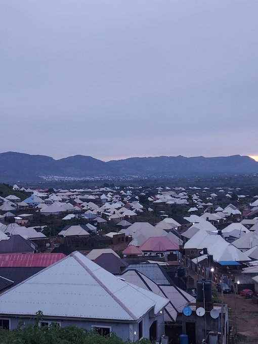 The image shows a view of a small town with numerous white buildings situated at the foot of a mountain range. The sky above appears cloudy, creating an overcast atmosphere. There are also several tents scattered throughout the scene, adding to the sense of a community gathering or event taking place in this area.