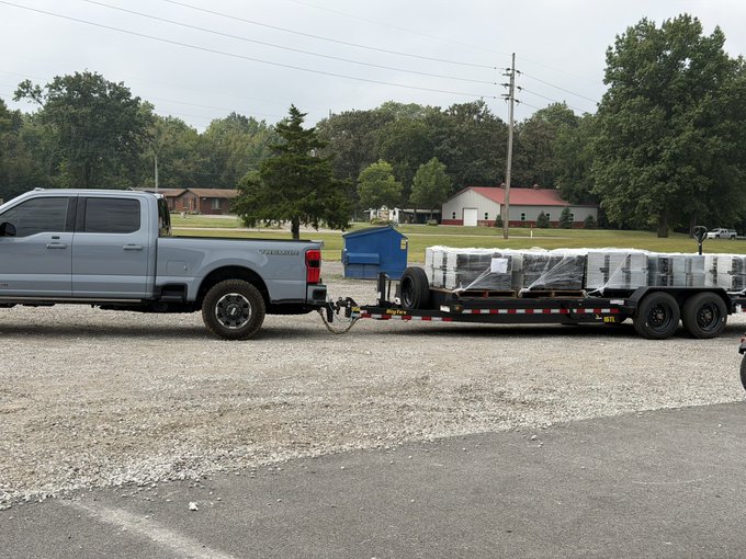 A light blue Chevrolet Silverado pickup truck parked on a gravel lot, hitched to a flatbed trailer loaded with multiple pallets of wrapped white bags, with trees and buildings visible in the background.