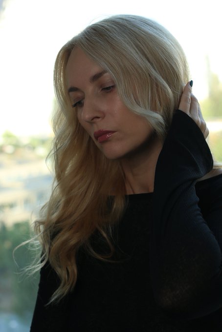 A portrait of a blonde woman wearing a black top, looking down thoughtfully with her hand in her hair against a soft outdoor background.