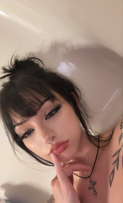 A young woman with dark hair styled in a messy updo poses thoughtfully with her finger to her lips, wearing a cross necklace and showcasing a delicate tattoo on her shoulder, embodying the mood behind the caption "baf."