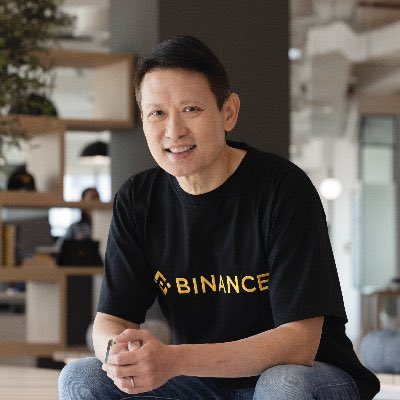 A man wearing a black 'BINANCE' t-shirt sits in a casual indoor setting, smiling at the camera with his hands clasped.