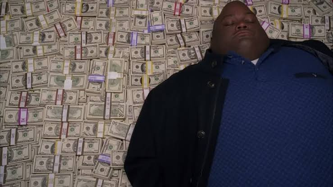 A man wearing a blue textured shirt and dark coat stands in front of a wall covered in banded stacks of US currency.
