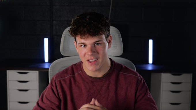 The image features a young man wearing a red sweater sitting at a desk with two white drawers behind him. He appears to be making a funny face or showing off his teeth while looking directly into the camera, possibly posing for a video. The background of the image is dark and out of focus, drawing attention to the subject in the foreground. There are also two blue lights present in the scene, which could potentially be part of the setup for filming the video.