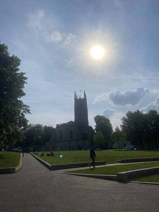 A bright sunny day shows a tall, dark stone church or cathedral tower rising against a blue sky with scattered clouds. The sun shines intensely overhead while people relax on the green lawn in the foreground park.