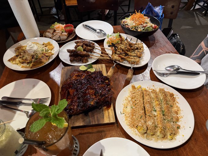 A table filled with an abundant feast of grilled meats, skewers, a large rack of ribs on a cutting board, and side plates of food. This aligns with the caption about "Gate's dinner, absolutely amazing," showcasing the impressive spread from the #GateEvent.