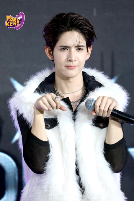 A young male performer on stage with dark hair, wearing a white fuzzy vest and black mesh sleeves, holding a microphone and making a heart gesture with his fingers; a 'PAPER KRAFT' logo appears in the corner.