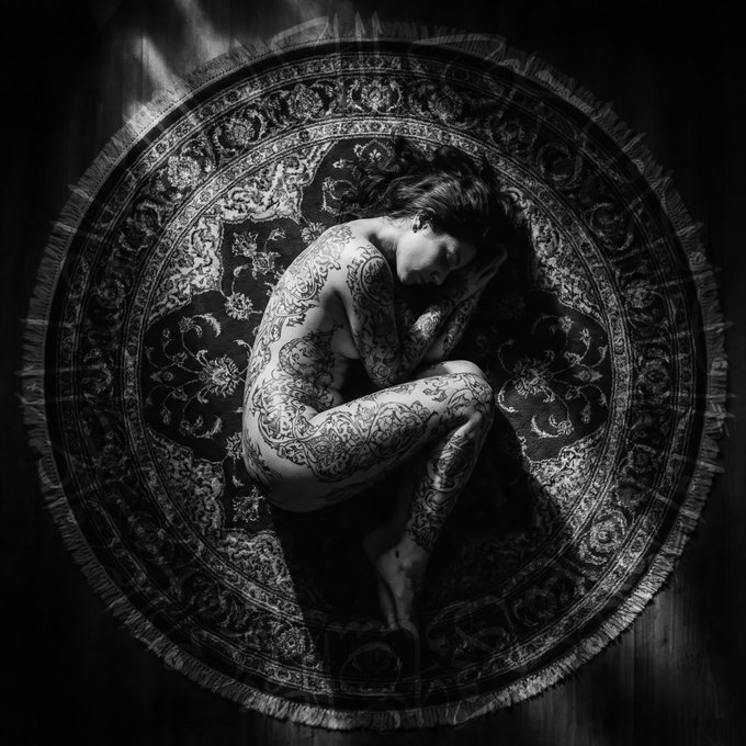 A black-and-white photograph of a tattooed figure curled into a fetal pose at the center of an ornate circular Persian rug, embodying the post's "Show me your dark side 'Circle'" theme through the visual harmony between the body's form and the rug's round design.