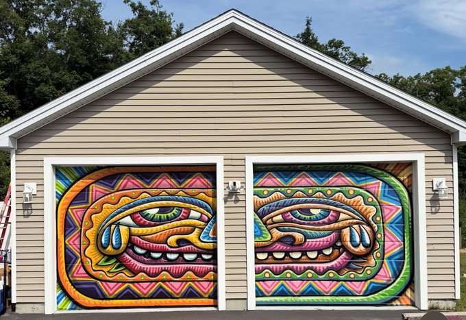 A house with two garage doors painted as a colorful mural forming two large, ornate tribal-style faces across the panels.