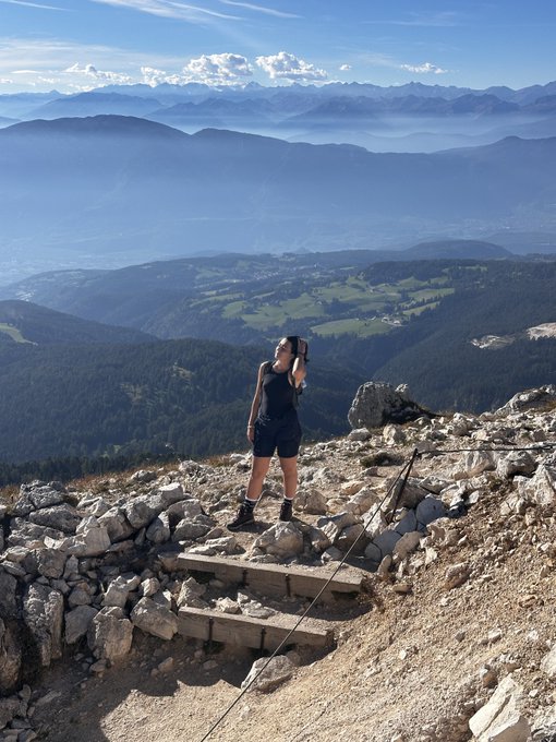 A hiker stands on rocky terrain at a mountain summit, using binoculars to view the distant valley and mountain ranges below.