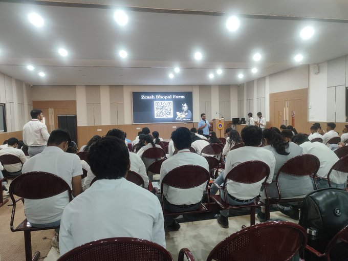 An audience of students in white uniforms sits in a large hall attending a lecture, with a presenter standing near a screen displaying a 'Zcash Bhopal Form' and a QR code.