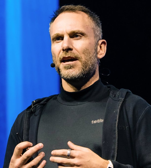 A man with a beard, wearing a black turtleneck with 'tet...' visible, speaks on stage with a microphone headset against a blue background.