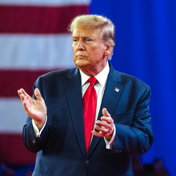 Donald Trump speaks with hands clasped, as he comments on the growing use of Bitcoin and cryptocurrency. The image shows Trump in a suit with a red tie, standing before an American flag backdrop.