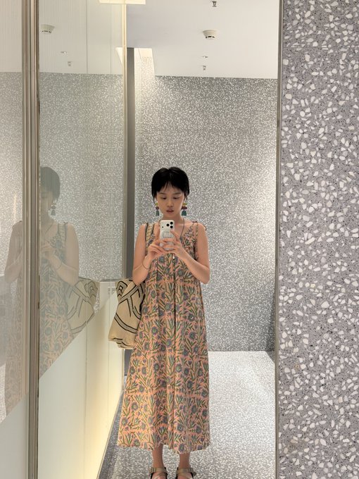 A young woman takes a mirror selfie in a glittery bathroom fitting room, wearing a patterned orange floral sundress with a beige handbag and statement earrings. The image appears unrelated to the post's content about a packed schedule of pickleball and travel to Singapore for Token2049, though her tired-but-styled look subtly echoes the caption's "no pressure, no gain" sentiment of pushing through exhaustion.
