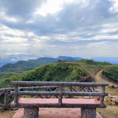 A wooden bench sits at a scenic hilltop overlook in the Chattogram hills, with a winding dirt trail leading toward distant mountain peaks under a dramatic cloudy sky, perfectly setting the stage for the hiker's journey toward their next summit, Keokradong in Bandarban.