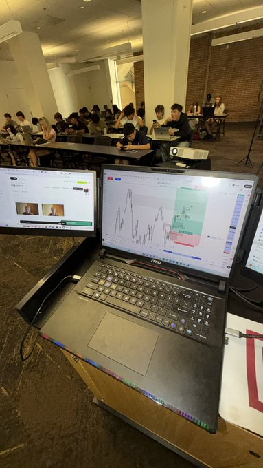 A laptop displays live trading charts and an active trading interface, with a classroom of students visible in the background working on their own computers. The image illustrates the caption's humor about teaching daytrading in real-time while half the students aren't paying attention, highlighting why most people struggle with the skill.