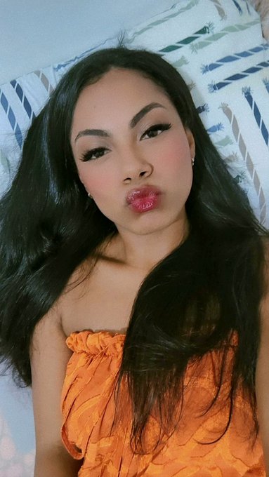 A portrait of a young woman with long dark hair, wearing an orange strapless top and posing with puckered lips against a striped background.
