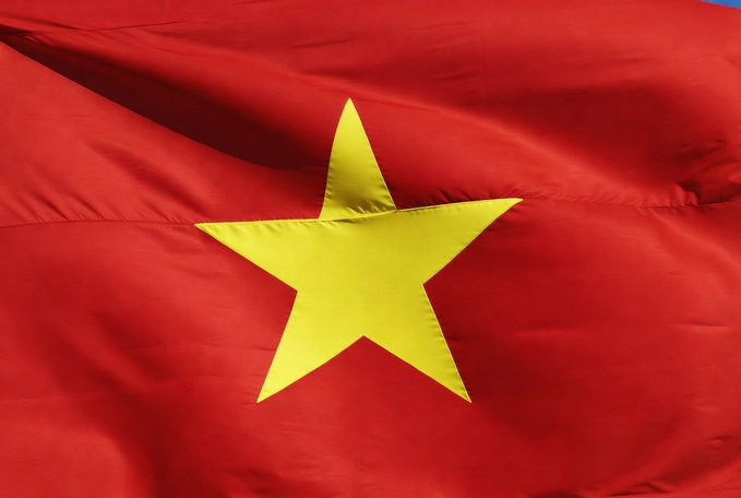 The image shows the Vietnamese flag waving, with its iconic red field and central yellow star. The flag visually represents Vietnam, the country referenced in the article about new penalties for unauthorized cryptocurrency trading.