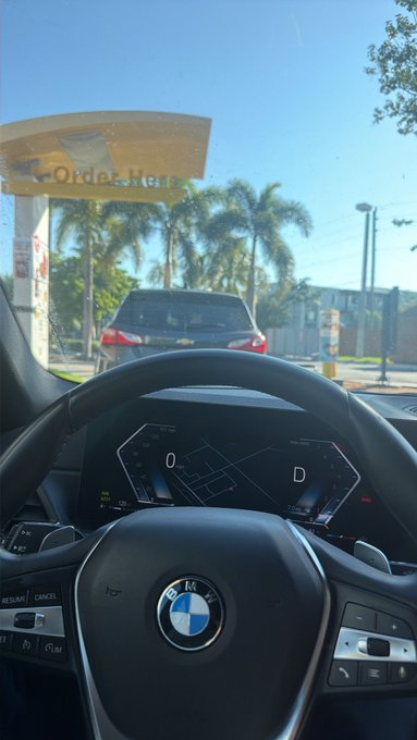 From the driver's seat of a BMW, a person waits in the drive-thru line at "ORDER HERE" under sunny blue skies, ready to fuel up before tackling a winning day on the markets.