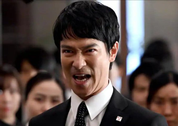 A man in a suit with a small pin on his lapel, appearing to yell or speak emphatically in what looks like a meeting or conference setting with people seated behind him.