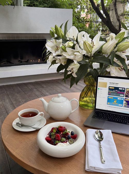 A serene outdoor setup featuring a wooden table with a white teapot, a teacup, a bowl of fresh berries, a laptop, and a vase of white lilies, all set against a backdrop of a modern fireplace.