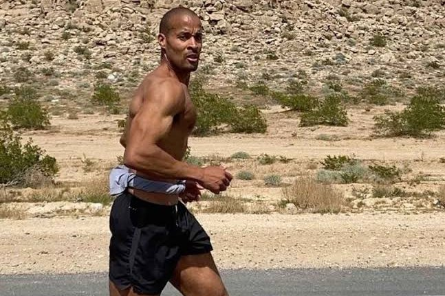 A muscular, shirtless man in black running shorts jogging through a desert-like landscape with rocky hills in the background.