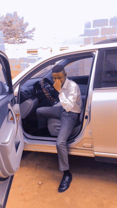 A man in a white dress shirt and gray pants sits in the driver's seat of a silver car with the door open, appearing contemplative with his hand over his mouth.