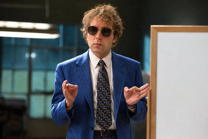 A man with curly hair wearing dark aviator sunglasses, a blue pinstripe suit, and a patterned tie stands in an office setting while gesturing with his hands.