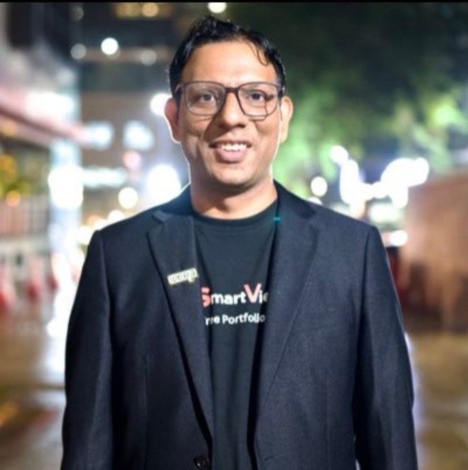A smiling man wearing glasses, a black t-shirt with 'Smart Vision Portfolio' text, and a black blazer, photographed outdoors at night.