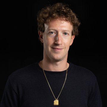 A curly-haired man with a subtle smile wears a black shirt and a gold pendant necklace against a black backdrop, visually representing Mark Zuckerberg as he addresses concerns about a potential AI bubble, acknowledging a "boom" in the industry on the Sources podcast.