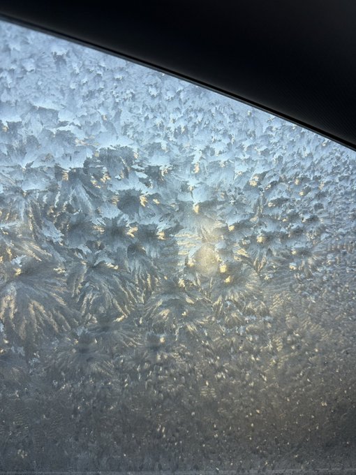 A car's window is completely covered in intricate, frost-like ice crystals that sparkle in the sunlight, illustrating the harsh winter conditions experienced in Canada.
