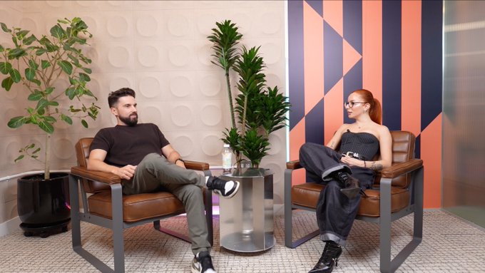 Two people sit in brown leather chairs during a podcast interview, with the man wearing a black shirt and the woman wearing a "GAP" shirt and glasses, set against a backdrop featuring plants and geometric orange and navy patterns. The image captures a casual, professional discussion setting that aligns with the post about a crypto-focused conversation featuring Brent Fulfer discussing institutional adoption and emerging markets.