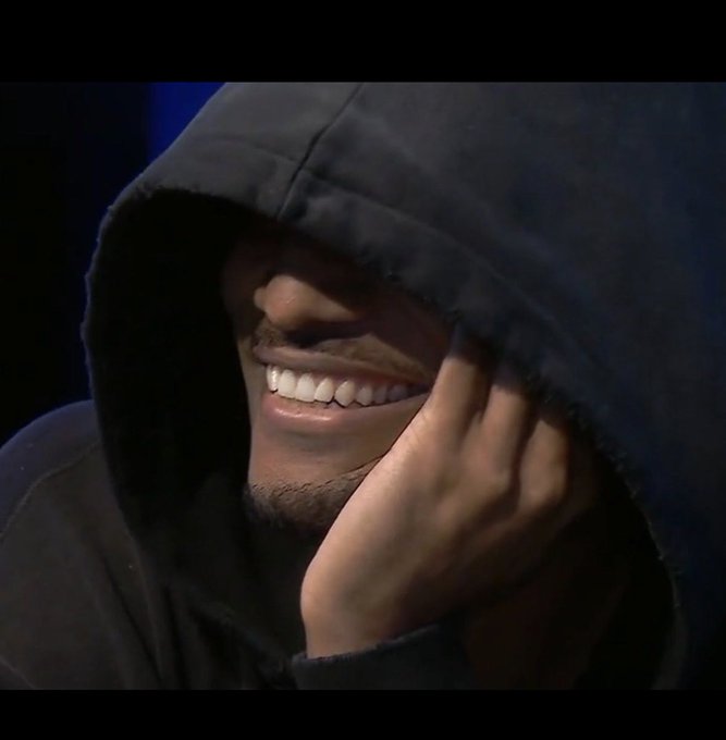 A hooded figure conceals his identity while smiling broadly, his face partially obscured by a dark hood and his hand resting near his cheek, conveying a sense of mystery and confident secrecy that mirrors the cryptic, cult-like hype surrounding the $DEALER memecoin comparison to the rise of $WHITEWHALE.