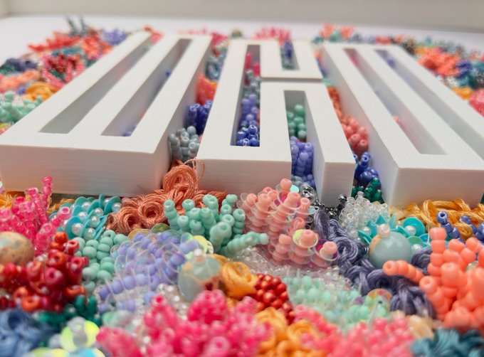 A close-up of a white 3D-printed 'OH' sign resting among a dense cluster of colorful beaded crafts, including clusters of pink, green, blue, and orange beads.