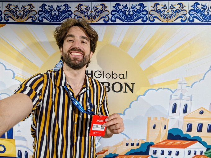 A smiling bearded man wearing a striped shirt and holding up a red 'JUDGE' badge for 'HUGO PORTEIRO', standing in front of a colorful illustrated backdrop reading 'Global Lisbon'.