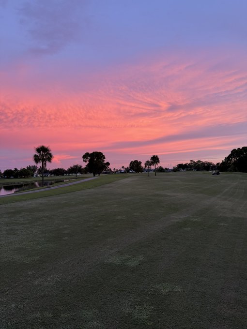 A scenic photograph of a golf course at sunset, featuring a vibrant pink and purple sky over green fairways, palm trees, and a small pond.