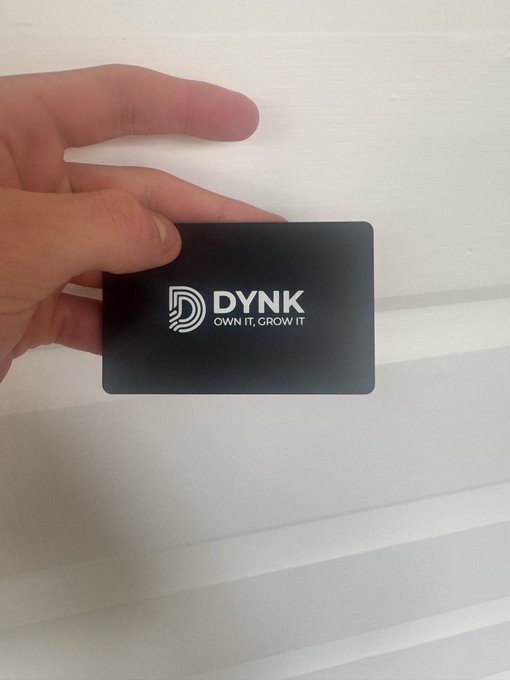 A hand holds up a black card with the text 'DYNK OWN IT, GROW IT' and a logo against a white slatted background.