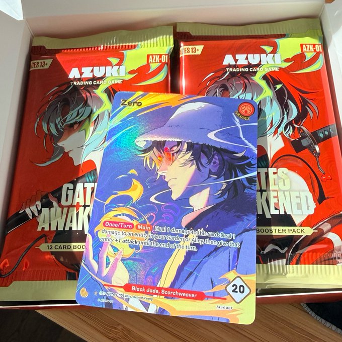 Two "AZUKI TRADING CARD GAME" booster packs (AZK-01) and a holographic promo card featuring "Zero" (Black Jade, Scorchweaver, 20 Power) displayed from a freshly opened box at the AZUKI Series 01 launch event.
