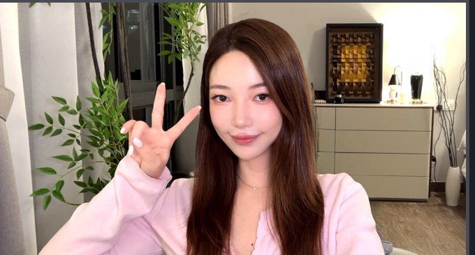 A young woman with long brown hair wearing a pink top flashes a peace sign while taking a selfie in a softly lit room with plants and decorative furniture in the background. The caption reveals she snapped the photo just before recording "Blockchain 100," teasing followers to guess her guest for a chance to win a cat picture.