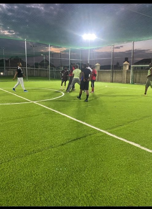 People playing football/soccer on an artificial turf field at night under floodlights, enclosed by netting.