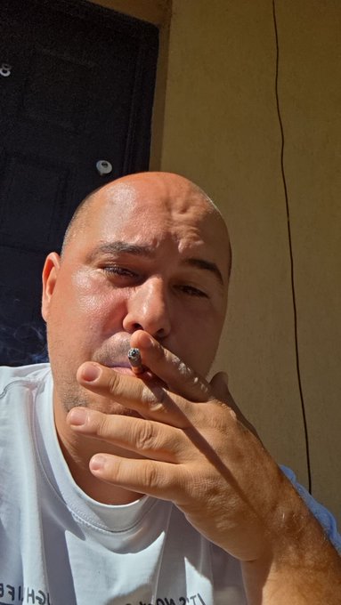 A bald man in a white t-shirt smokes a cigarette while taking a close-up selfie, with a dark mesh surface and beige wall visible in the background.
