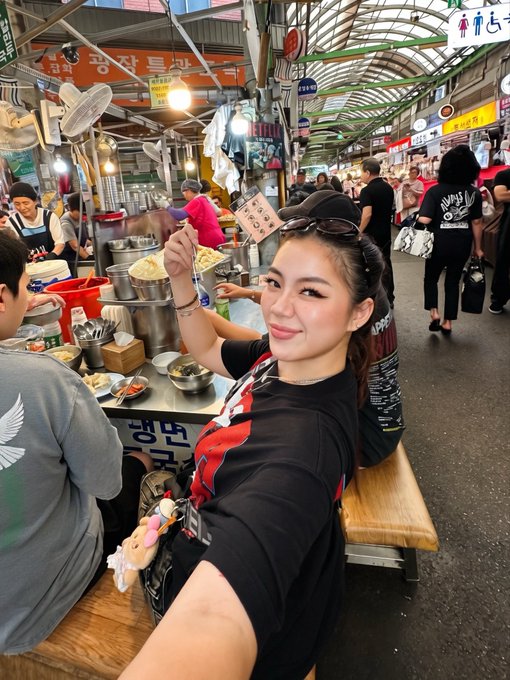 A selfie taken by a young woman in a black t-shirt sitting at a food stall in what appears to be a bustling Korean market, with Korean signage visible in the background.