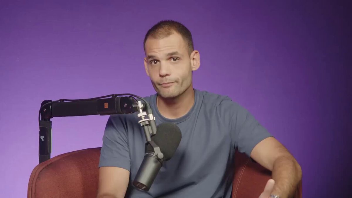 The image shows a man sitting in a chair with his hands together, wearing a blue shirt. He is holding a microphone close to his face, suggesting that he might be participating in an interview or speaking event. The background of the image is purple, which could indicate a stage setting for the event taking place.